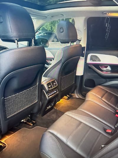 The image shows the interior of a modern car, likely a luxury SUV, with black leather seats and a panoramic sunroof. The view is from the back seat, looking forward. There are visible air vents, a door panel with ambient lighting, and seat pockets. The flooring appears to be a dark carpet. There are no people or animals visible in the image. The exterior scene visible through the windows appears to be a sunny day with trees and other vehicles in the background, suggesting an outdoor parking area. There are no specific landmarks or indicators that would pinpoint this location to Moscow, Russia.