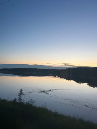 The image depicts a calm body of water, likely a lake or river, reflecting a serene sky at dusk. The sky transitions from a pale blue at the top to softer hues of yellow and orange near the horizon, indicating the setting or rising sun. A dark silhouette of a dense forest lines the far bank of the water. The water's surface is smooth, creating a near-perfect mirror image of the sky and the treeline. In the foreground, the blurred dark green of vegetation suggests the viewer is positioned on the shore or in a vehicle moving along the water's edge. The scene evokes a sense of tranquility. The provided information about the location being "Суда, Russia" is not visually verifiable from the image itself; the image shows a natural landscape without any specific identifiable landmarks of the city.