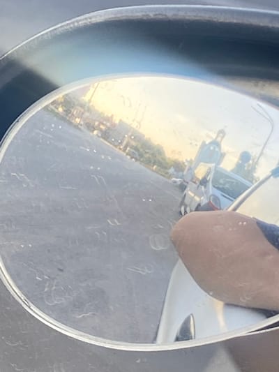 The image is a close-up, distorted reflection in a curved mirror, likely a side-view mirror of a vehicle. The primary subject is a portion of a person's arm, its skin appearing tan and slightly textured, with what look like small water droplets or dust particles on the surface. This arm is positioned in the upper right of the frame, partially obscuring the reflected scene.
The reflection itself shows a bright, outdoor setting, suggesting it is daytime, possibly late afternoon or early evening due to the warm, golden light. The scene reflected in the mirror is blurry and upside down. In the background, there appears to be a white vehicle, possibly a car or small van, with some metallic parts visible. Further beyond the vehicle, a town or city landscape is visible, with buildings and trees lining a street. The sky in the reflection is bright and appears to be a clear or lightly clouded sky. The ground visible in the reflection seems to be asphalt or a similar paved surface.
The overall impression is a snapshot of a moment, perhaps during a car journey or while the vehicle is parked. The water droplets on the arm and the reflection suggest potential recent rain or washing. The location context of Pavlohrad, Ukraine, does not provide specific visual cues within the image itself, as the reflected scene is generic. There is no visible text in the image. The mood is neutral, a simple capture of a reflective surface and its surroundings.