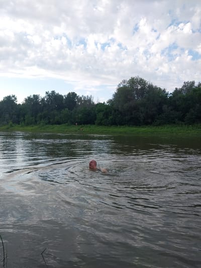 A middle-aged bald man with his mouth open is swimming in a wide, murky river. He is partially submerged, with his head and shoulders visible above the rippling water. His eyes are closed, and he appears to be exerting himself. Green vegetation lines the opposite bank, with a dense forest beyond. The sky is overcast with scattered white clouds. The scene is likely a natural body of water within or near the city of Chkalov, Russia, possibly a park or recreational area. The water is calm with some circular ripples around the swimmer. There are no other individuals or objects in the immediate vicinity of the man in the water.