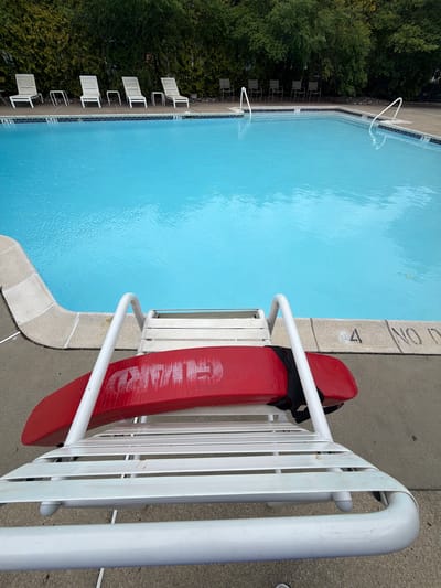 This is an outdoor scene at a swimming pool in Edison, New Jersey, USA. A red lifeguard rescue buoy is hanging on a white lifeguard chair at the edge of the pool. The pool water is clear and blue. Along one side of the pool are several white lounge chairs. Green trees and bushes line the area behind the lounge chairs. Pool depth markers are visible.