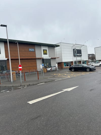 The image depicts an outdoor scene in Aberdeen City, United Kingdom. It features a McDonald's restaurant and a PureGym fitness center. A black car is parked beside the PureGym building. A large white directional arrow is painted on the asphalt roadway. A no entry sign is visible near the entrance of the McDonald's. There are other parked cars visible in the background. The sky is overcast, suggesting a cloudy day. Birds are flying near the top of the frame.
