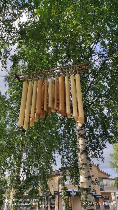 The image shows a view looking up through the branches of a birch tree in Iisalmi, Finland. Suspended from a metal rack above the tree branches is a collection of wooden tubes of varying lengths and thicknesses, resembling a wind chime or a musical installation. The tubes are light brown and cream in color. In the background, behind the tree, are buildings that appear to be shops or commercial establishments, with signs visible that read "Puistelli." A pedestrian crossing sign is also visible. The sky is partly cloudy. The date stamp "2026/7/8 18:45" is visible in the lower right corner.