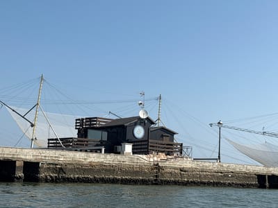 This is a photograph of a fishing hut located in Cesenatico, Italy, specifically along the canal port. The hut is dark wood and appears to be elevated on a concrete structure that extends into the water. There are large fishing nets with poles extending from the hut and into the sky, indicating active fishing operations. A satellite dish is mounted on the roof of the hut, along with an antenna. A circular decorative emblem, resembling a compass rose, is visible on the side of the hut. The water in the foreground is rippled and reflects the blue sky. The overall scene is bathed in natural sunlight under a clear blue sky.