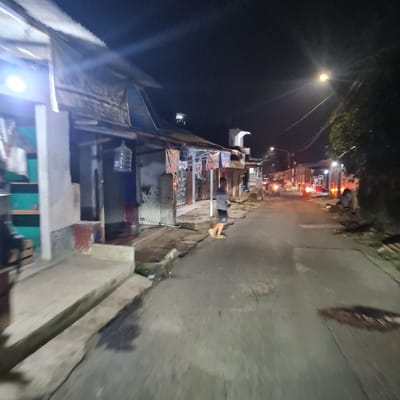 The image shows a narrow street in Kecamatan Tamansari, Indonesia, at night. The street is lined with small, single-story buildings. One building has a large water cooler on the side. A single person walks away from the camera, down the street. The street is paved with asphalt and appears to be lit by a streetlamp in the distance. The environment is dimly lit, with the buildings casting long shadows. There are a few parked vehicles in the distance. The atmosphere is quiet and peaceful.  The scene appears to have been taken in a residential neighborhood, with minimal traffic.