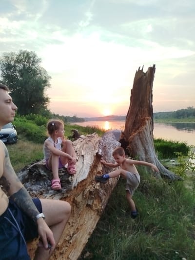 In Topolina, Poland, at sunset, a man and two children are by a river. The man, partially visible on the left, has tattoos on his arm. A young girl with blonde pigtails sits on a large fallen log. A boy is actively climbing on the log. The river flows behind them, with trees and vegetation on the far bank. A car is parked behind them on the grass. The sky is cloudy with the sun setting in the distance, casting a warm glow on the scene.