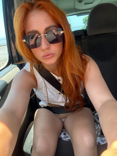 A young woman with long, bright red hair is sitting in the passenger seat of a car. She is wearing large, dark sunglasses and a white top with eyelet details and a gold chain. A black seatbelt is visible across her chest. Her lips are pursed. The interior of the car is visible, including the back of the driver's seat and headrest. Through the window, a glimpse of the outdoors can be seen, with a road and some trees, suggesting a location that could be along a scenic route, potentially near a coastal area given the bright light and expansive sky. This scene is likely taking place in or around the city of Novoselje, Montenegro.