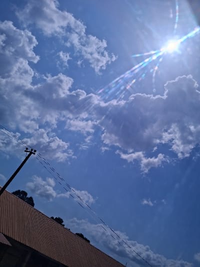 The image depicts a bright, sunny daytime sky over Takum, Nigeria, with a clear blue expanse partially obscured by scattered cumulus clouds of varying density. The sun, a brilliant white orb, is prominently positioned in the upper right quadrant, casting strong, multicolored lens flares that radiate downwards and to the left. In the lower portion of the frame, the upper section of a building with a light brown, possibly corrugated or tiled roof runs horizontally, slightly angled upwards from left to right. Above the building's roofline, dark green tree foliage is visible against the sky. A dark utility pole, likely wooden, extends diagonally upwards from the lower left corner, bearing multiple thin electrical or communication wires that stretch horizontally across the mid-left area of the sky. No individuals, vehicles, or specific actions are discernible.