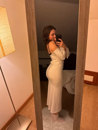 An indoor scene depicts a female individual, with medium-length dark brown hair, taking a mirror selfie. She is positioned to her right side, facing a full-length mirror with a light brown frame. She wears a light-colored, form-fitting off-the-shoulder garment that extends below the knee, revealing a black bra strap on her left shoulder. She holds a dark-colored smartphone in her left hand, pointing it towards her reflection, and her right hand touches her chin/mouth area as she displays a subtle smile. Her feet, clad in white translucent socks, rest on a white fluffy rug positioned on a light-colored wooden floor. To the immediate left of the mirror, a floor lamp with an orange rectangular fabric shade is visible. The reflection in the mirror's background shows a dimly lit room with a warm ambient light, featuring a dark furniture piece, possibly a bed or dresser, partially covered by a light-colored textile. The specific location within the city of Cava de' Tirreni, Italy, cannot be determined from the provided visual information.