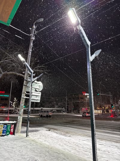 A snowy urban street scene at night. Snowflakes swirl in the dark sky, illuminated by a bright street lamp on the left. A snow-covered sidewalk runs along the right edge. In the distance, traffic lights and a building with a red sign are visible. Utility poles and wires crisscross the sky.