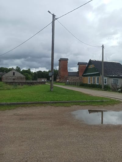 The image depicts a rural scene in the Крэўскі сельскі Савет, Belarus, under a cloudy sky. A gravel road runs through the foreground, with a large puddle reflecting the overcast sky and a utility pole. To the left of the road, a grassy area is bordered by a low stone wall and a building constructed from stone and light-colored brick. Beyond this building, trees form a backdrop. To the right of the road, a house with green and light-colored wooden siding and a dark, sloped roof stands prominently. Adjacent to the house are brick ruins, with arched openings and dark, sloped roofs. A second, smaller utility pole is visible near the house. The overall impression is one of a historic or agricultural area in a state of transition or preservation.
