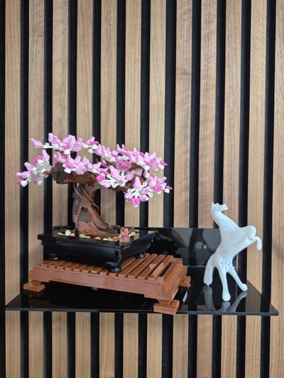 A Lego cherry blossom bonsai tree is displayed on a shelf in front of a wooden slatted wall. The tree has a brown trunk and branches with many pink and white blossoms. It sits in a black pot filled with small pebbles and a tiny pink duck figurine. The pot is elevated on a brown Lego platform. To the right of the bonsai, a white abstract sculpture resembling a horse is placed on the same shelf. The shelf is black and reflective, showing a faint reflection of the horse sculpture. The background is a wall composed of vertical wooden slats alternating with dark gaps. The scene appears to be an indoor display, possibly in a home or a retail space in Barbing, Germany. There are no people or actions depicted in the image.