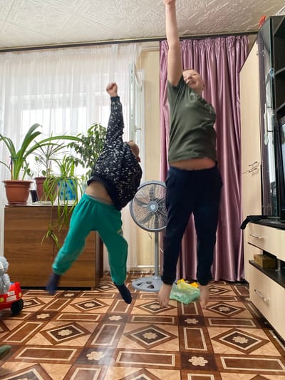 The image shows two individuals posing in what appears to be a domestic interior. The location may or may not be Bely Klyuch, Russia.
The upper figure is an adult male wearing a green shirt and dark pants, with his arms outstretched. He is positioned horizontally in the frame. A fan can be seen nearby.
The lower figure appears to be a child in a black and white sweater and green pants, also with arms outstretched, posed in a similar horizontal fashion. Several potted plants are visible near the child.
The background features curtains, furniture, patterned flooring, and textured walls.