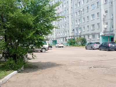In a sunny daytime scene in Arseniev, Russia, a residential apartment building with a light grey facade dominates the background. The building has multiple stories, each with numerous windows and air conditioning units visible. In the foreground, a paved area serves as a parking lot with several cars parked. To the left, a large, leafy green tree provides shade, with its branches extending into the frame. The ground is a mix of asphalt and dirt, showing signs of wear and tear. In the middle ground, a white van and a white minivan are parked. Further to the right, a silver hatchback and a dark grey hatchback are also parked. The sky is bright and clear, suggesting good weather. There is no visible text in the image.