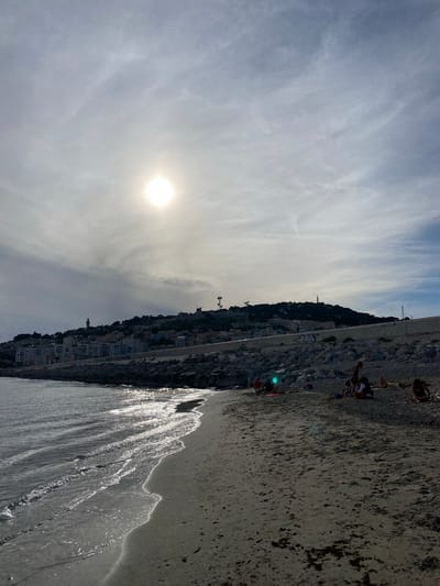 The image captures a coastal scene in Sète, France, under a bright sun obscured by thin clouds, likely in the late afternoon. The scene is dominated by the Mediterranean Sea on the left, a dark pebbly beach in the center and right foreground, and Mont Saint-Clair in the background.
The sky is partly cloudy, with the bright, white-yellow sun positioned in the upper-left quadrant, creating a strong glare and lens flare. Mont Saint-Clair, a hill, is densely covered with light-colored buildings and trees. A distinctive white cylindrical lighthouse stands prominently on the left slope of the hill, while multiple communication towers with antennae are visible on the hill's crest towards the right.
A concrete retaining wall, a light grey color with some darker patches and graffiti near the center-right, runs horizontally along the base of the hill. Below this wall, a substantial barrier of large, irregular grey rocks extends along the shore.
The sea, on the left, shows gentle waves breaking onto the beach. The water's surface intensely reflects the sunlight, creating shimmering, elongated streaks. The beach itself is composed of dark grey, coarse sand or small pebbles, with scattered patches of darker debris, possibly seaweed.
Approximately ten individuals are present on the beach, engaged in leisure activities. On the left side of the beach, near the rock barrier, a group of three to four people are seated or resting; one individual exhibits a notable greenish-blue light reflection on their back. Further to the right, a second group of three to four individuals, including at least one adult and a child, are seated on the sand, some on towels. Towards the far right, two additional individuals are visible, one lying prone and another seated nearby. There are no discernible active interactions between the individuals or groups; they appear to be relaxing independently.