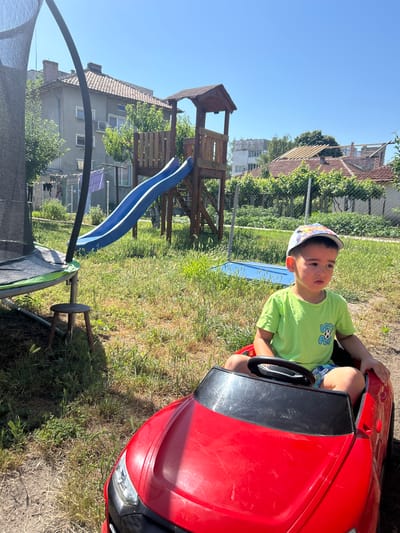 A young boy wearing a green t-shirt and a colorful cap sits in a red toy car in a backyard. The backyard features a trampoline with a black net and a blue slide attached to a wooden play structure. There are houses and buildings in the background. The image was taken in Belene, Bulgaria.