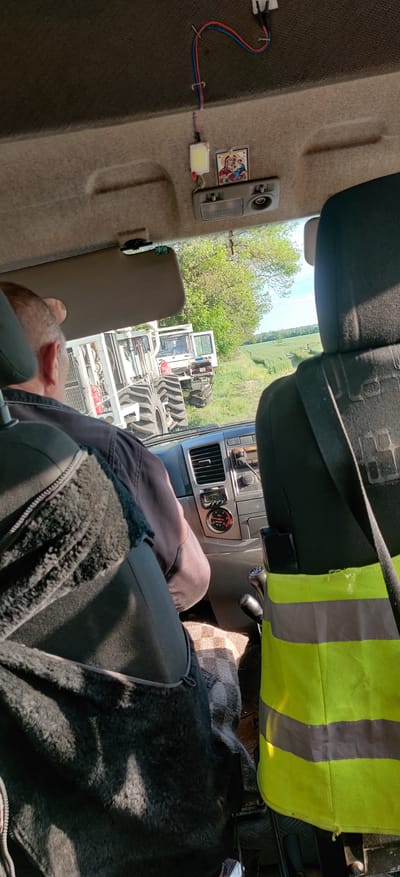 The image shows the interior of a vehicle, possibly a tractor, parked in a field near Randava, Ukraine. A man is seated, partially obscured by the seat, wearing a black jacket with a sheepskin-like lining. A high-visibility vest is draped over the passenger seat. Visible through the window is another piece of machinery, appearing to be a tractor or agricultural equipment. The vehicle's dashboard includes a digital display showing the time as 12:18. The vehicle's interior includes a light fixture and an attached sticker. A red and blue wire is attached to the fixture. The surrounding environment features green fields and trees.