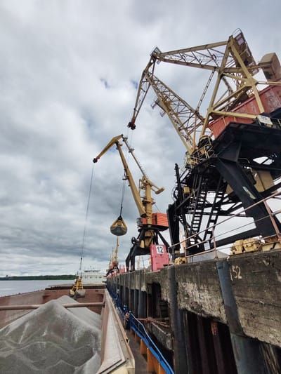 The image depicts a harbor scene in Rybinsk, Russia, under a cloudy sky. Two large, yellow cranes dominate the foreground, positioned on a concrete pier. The crane on the right has black supports and a number "12" painted on its structure. A loaded bucket hangs from the crane in the center. Below the cranes, a ship's hold is visible, filled with piles of gravel or similar material. In the distance, a white ship is docked. The pier is weathered and marked with the number "152".