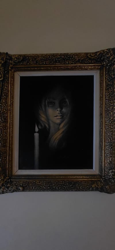 A framed painting of a woman's face illuminated by a single candle hangs on a wall. The painting is in a dark, dramatic style, with the woman's face emerging from shadow. A lit candle is visible to the left of the woman's face, casting a soft glow on her features. The painting is encased in an ornate, gold-colored frame. The wall itself is a neutral, light color. This scene is located within a residential setting in the Municipal Unit of Aegio, Greece.