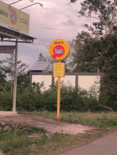 This image depicts a roadside scene in Bandasafa, Indonesia, featuring two prominent signs related to a telecommunications provider. In the foreground, a paved path or road is visible, partially covered by overgrown grass and dirt, leading off to the right.

The central focus is a tall, yellow pole supporting two signs. The upper sign is round and bright yellow with a red circular shape inside, prominently displaying the text "Mitra im3". Below it, a smaller rectangular yellow sign with black text provides additional, albeit unreadable, information.

To the left, a larger billboard is partially visible against the cloudy sky. It also features "Mitra im3" at the top and a partially readable company name, "PT.KOTAJANTHO," below. The billboard is supported by metal beams and posts.

The background is dominated by dense, green foliage and trees, suggesting a rural or semi-urban setting. Behind the central signs and trees, a white wall or fence-like structure is visible, behind which a small building with a corrugated metal roof can be seen.

The sky is overcast with diffused light, indicating either an early morning, late afternoon, or simply a cloudy day. There are no people visible, and the scene appears static. The signs suggest a point of sale or information center for the "im3" mobile operator, which is common in Indonesia, fitting the provided location context.