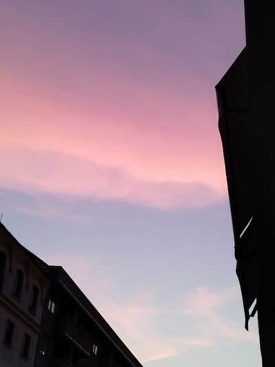 This image shows a twilight sky with soft pink and purple hues over buildings in Jerez, Spain. The sky is a gradient of color, with lighter shades near the horizon and deeper purples higher up. Wispy clouds are scattered across the sky, catching the light and adding texture. In the foreground, silhouetted buildings are visible against the colorful sky. One building is dark and angular, with a sharp edge cutting into the frame. Another building to the left is lighter in tone and shows architectural details such as arched windows and balconies. The scene appears to be a city landscape at dusk or dawn, with the vibrant sky providing a dramatic backdrop to the urban environment.