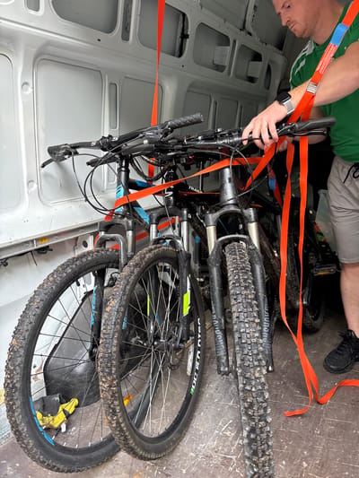 A man is standing in a van, securing two dirt bikes with orange straps. The man, wearing a green t-shirt and grey shorts, is holding the handlebars of one of the bikes. The bikes are black and blue and have mud on their tires. The van is white and appears to be parked indoors. The scene is illuminated by natural light coming from outside the van. The location is not specified in the image, but if the city of Birkenau, Germany, were added as a context, it would be a detail outside the visual information.