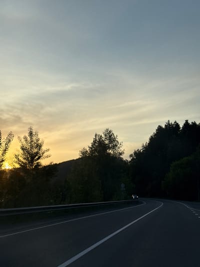 The image depicts a winding asphalt road at either sunrise or sunset in Manzherok, Russia. The sky transitions from a bright, warm yellow-orange at the horizon on the left, where the sun is partially visible behind silhouetted trees, to a cooler light blue-grey in the upper portion, with wispy clouds. Dark, forested hills form the distant background. Along the left side of the road, a metallic guardrail is present. The road features a solid white line on the left and a dashed white center line. A vehicle with illuminated headlights is visible in the distance on the right side of the road, navigating the curve. The immediate environment surrounding the road consists of dense, dark green trees and foliage, appearing silhouetted against the bright sky on the left and in shadow on the right. No individuals are visible.