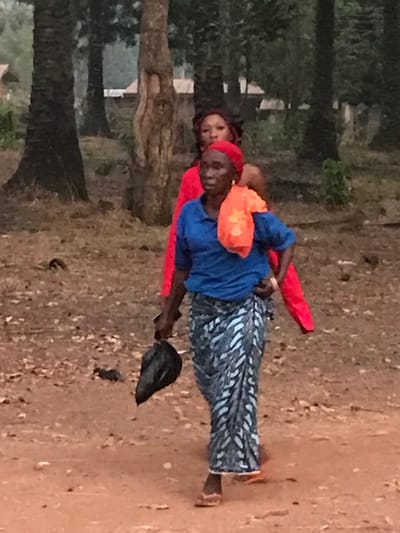 A woman wearing a red headdress carries another woman on her back. The woman being carried also wears a red top. The carrier has on a blue shirt and a blue and white patterned skirt. She has an orange bag draped over her neck and holds a black object. They are on a dirt path with scattered leaves. Trees are visible in the background.