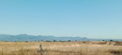The image captures a wide, horizontal landscape under a clear, pale blue sky. In the foreground, a blurry wire fence runs horizontally across the frame, suggesting the perspective of a moving vehicle. Beyond the fence, a dry, golden field stretches out, with sparse vegetation and the impression of tilled soil in curved patterns on rolling hills. A tripod-like object is visible in the left mid-ground. In the middle ground, a line of scattered trees and shrubs dots the landscape. The background is dominated by a range of hazy, blue mountains that extend across the horizon. The overall scene depicts a rural, possibly agricultural area, with a sense of dry weather and open space. The image is consistent with a view of the countryside in Quismondo, Spain, known for its agricultural lands and proximity to mountainous terrain.