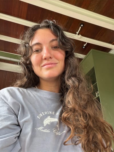 A young woman with curly brown hair, styled with gray highlights, smiles gently at the camera. She's wearing a light blue long-sleeved shirt adorned with the words "Sunshine Beach, Malibu, California" encircling a drawing of a dolphin. The background shows a wooden ceiling, white beams, and a light green cabinet partially visible. The image is taken at a slightly tilted angle, creating an interesting perspective.