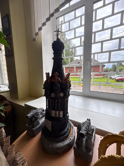 The image shows a dark, ornate, cathedral-like sculpture with a cross on top, placed on a wooden table next to a window. Two smaller, grey sculptures, one resembling a cannon and the other a sheaf of wheat, are positioned on either side of the larger sculpture. The window has a decorative grid and looks out onto a street with red brick buildings, green grass, parked cars, and trees under a cloudy sky. The overall setting appears to be indoors, with elements of decoration and display. The image does not contain any people or any specific identifiable information about the location within Ветрынскі сельскі Савет, Belarus.