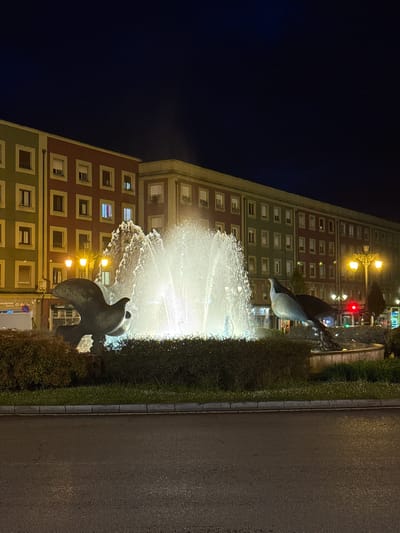 The image depicts the Uria Street fountain, a notable landmark in Oviedo, Spain, during nighttime. The fountain features bronze statues of dolphins, with water jets illuminating the scene. In the background, a large building with multiple windows, displaying a combination of red, green, and beige colors, is visible. Streetlights cast a warm glow, adding to the urban ambiance. A sidewalk and a street are observed on the right side of the frame.