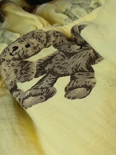 The image is a close-up, oblique, full shot of a light yellow, possibly cotton or linen, textured fabric with a repeating pattern of brown teddy bears. The fabric is folded and draped, creating shadows and highlighting the texture. The teddy bear pattern is depicted in a stylized, somewhat faded manner, with dark brown lines and shading against the lighter background. The bears are shown in various poses, some appearing to be sitting or lying down. The lighting is soft and diffused, casting subtle shadows on the fabric. The image is focused on the texture and pattern of the textile itself. There is no information in the image that would place it in Uchkun, Kyrgyzstan.