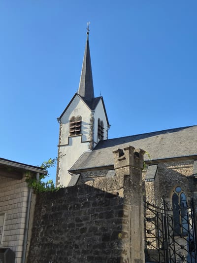 The image shows a detail of a church spire and adjacent building in Schengen, Luxembourg. The scene is viewed from a low angle, looking upwards against a clear blue sky. The church spire is tall and pointed, covered in grey shingles. Below the spire, a section of the church's white facade is visible, featuring a window with multiple narrow panes and a decorative stone surround. To the right of the spire, a section of a stone wall with a textured, dark grey surface is prominent, along with a portion of a building with a grey tiled roof and a white tiled section. Some greenery, likely ivy, is growing on the stone wall and near the roofline.
The lighting suggests it is daytime, likely late morning or early afternoon, given the strong shadows cast by the architectural elements. The weather appears to be clear and sunny. There is no text visible in the image. The image captures a static architectural scene, with no people or specific events taking place. The focus is on the interplay of light, shadow, and the textures of the stone and roofing materials.