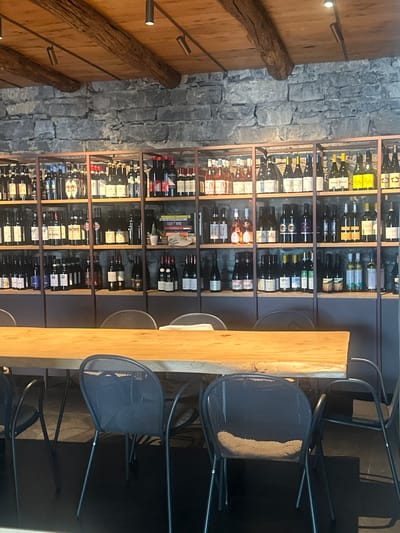 The image shows an interior scene of a wine cellar or tasting room with a rustic aesthetic. A long, live-edge wooden table is in the foreground, set with several dark metal mesh chairs. Behind the table, extensive shelving units are filled with numerous bottles of wine. The shelves are constructed with dark metal frames and wooden planks, and they are built against a rough stone wall. The ceiling features exposed wooden beams and dark, track lighting fixtures. The bottles on the shelves vary in color, labeling, and shape, indicating a diverse selection of wines. On the middle shelf, towards the right side, a few books are visible, including one with "EASY WINE" on its spine. The lighting in the room is dim, contributing to a cozy and sophisticated atmosphere. The overall impression is that of a well-stocked establishment designed for wine appreciation. This scene is located in Varenna, Italy.