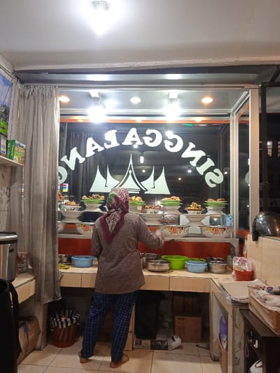 The image shows a woman standing in a small restaurant with a large glass storefront. The restaurant is located at the coordinates -7.558, 110.777 in Kecamatan Kartasura, Indonesia. The woman is wearing a brown sweater and a head covering. She is standing in front of a counter with several dishes displayed behind a glass partition, including a large, white sign with the word "Singga" written in a stylized, reverse font. The restaurant appears to be open for business and the woman may be preparing to serve customers. Behind her are more dishes, a large bin, and a partially visible box. A black helmet lies on the counter on the right side of the frame. The counter has tile flooring, and the walls are painted a pale orange. The storefront is lit by several overhead lights.
