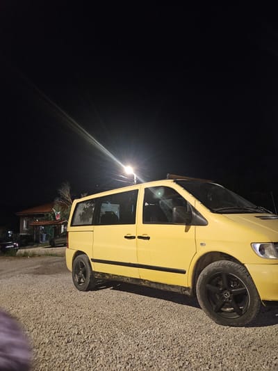 The image depicts a yellow Mercedes-Benz Vito van parked on a gravel surface at night. The van is positioned to the right of the frame, angled slightly towards the viewer. It has black rims and a row of aftermarket lights mounted on its roof. In the background, to the left, is a house with a red roof and a flag flying from its porch. A street light illuminates the scene from above and to the left, casting light rays across the dark sky. The overall environment appears to be rural, likely on the outskirts of Zlataritsa, Bulgaria, given the presence of the house and the unpaved ground. There are no people clearly visible in the frame, and the van appears to be stationary. The scene is captured from a slightly low angle, emphasizing the presence of the van.