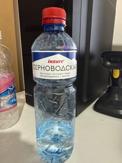 The image displays a close-up, eye-level view of a plastic bottle of water on a white surface, likely a desk or counter. The bottle is partially filled with clear blue liquid and has a red cap. The label on the bottle is predominantly blue and white, with red text that reads "inzare" and larger Cyrillic text below it. To the left of this bottle is another partially visible plastic bottle with a pink and blue label. In the background, a dark, upright object, possibly a computer tower, is present. To the right, a black scanner with a yellow button is visible. The lighting suggests an indoor environment. There are no people or animals in the image. The scene is static and does not depict any actions or interactions. The image does not provide any explicit or implicit information about the location in the city of Mayrtup, Russia.