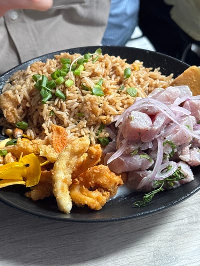 A close-up shot captures a vibrant Peruvian meal served in a black bowl on a light wooden table. The dish is a combination of several popular Peruvian elements.

On the left side of the bowl is a generous portion of brown, seasoned rice, likely an arroz chaufa-style fried rice, generously garnished with chopped green onions. Adjacent to the rice, towards the top, is a serving of ceviche, featuring chunks of white fish marinated and mixed with thinly sliced red onions and fresh herbs. A light, milky liquid, characteristic of leche de tigre, is visible at the bottom of the ceviche.

To the right side of the bowl, a pile of golden-brown fried seafood, resembling calamari or shrimp, adds a crispy texture to the plate. Below the fried seafood, several bright yellow plantain chips are visible, along with a few roasted corn kernels (cancha). A small, thin, brown chip, possibly a fried plantain or cassava chip, is placed at the very top edge of the bowl, serving as a garnish.

The lighting is bright and even, highlighting the colors and textures of the food. The background consists of a blurred person's arm and clothing (a beige sleeve and a blue denim-like fabric), suggesting an indoor setting, likely a restaurant or home in Jesús María, Peru. There is no visible text in the image.