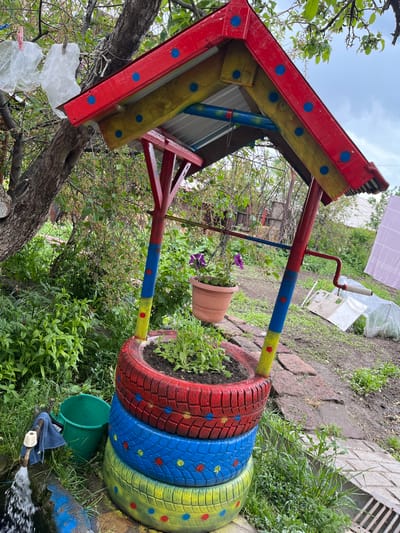 In Lusagyugh, Armenia, a brightly painted, makeshift wishing well stands in a garden. The structure, built from colorful tires stacked and painted red, yellow, and blue, is topped with a multi-colored roof. A terracotta pot with purple flowers hangs from a support beam, and a single plant grows from the top tire. Adjacent to the well, a green bucket and a faucet with water flowing into a blue-painted basin are visible. The surrounding area is overgrown with greenery, with trees overhead and a cloudy sky above. Laundry hangs on a clothesline in the background. The scene depicts a charming, rustic backyard decoration.