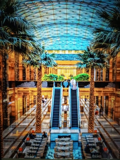 This image depicts the interior of a modern, multi-level building with a large, arched glass ceiling. The architecture features warm-toned wood paneling and reflective surfaces, giving it an opulent feel. Two prominent palm trees flank a central walkway. Escalators are visible on either side of the walkway, leading to upper levels. In the foreground, tables and chairs are set up, suggesting a dining or seating area. Several people are present, some walking and others seated at the tables. One group of people, two individuals in traditional attire, are on an escalator. The overall impression is of a well-maintained, upscale commercial or public space. The location is identified as Golbembal, Nigeria.