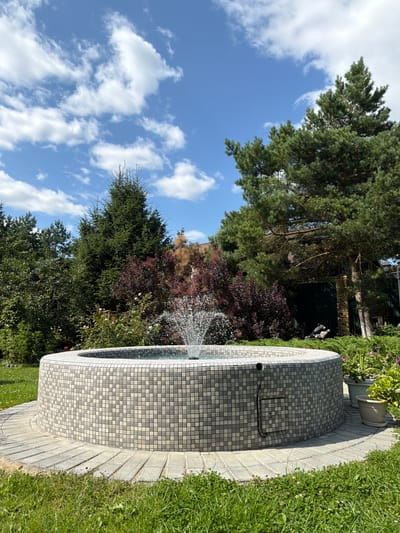 In the village of Zhostovo, Russia, a circular fountain, clad in a mosaic of small grey and beige tiles, stands in a well-maintained garden. Water sprays from the center of the fountain, creating a delicate mist. The fountain is surrounded by a paved walkway made of rectangular stones. Lush green grass carpets the foreground, with various trees and shrubs forming a dense backdrop. Tall evergreen trees, including what appear to be spruce and pine, dominate the landscape, interspersed with dark red foliage plants and flowering bushes. The sky above is a vibrant blue, dotted with fluffy white clouds, suggesting a clear, sunny day. The scene depicts a tranquil outdoor setting, likely a private garden or a public park space.
