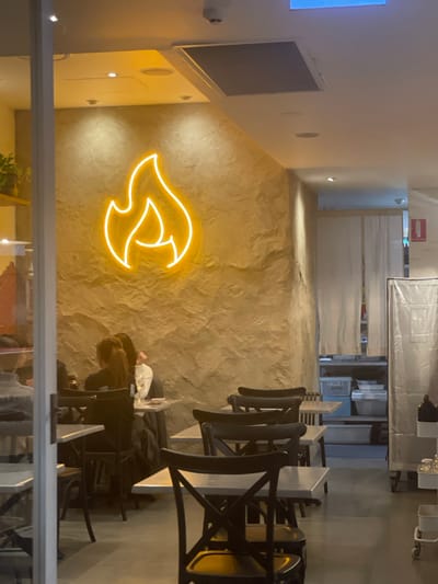 The image shows the interior of a restaurant. A neon sign shaped like a flame is visible on a textured, light brown wall. Several tables and chairs are arranged in the dining area, some occupied by diners. In the background, white curtains hang over a doorway, and some storage shelves are visible. The lighting is warm, primarily from overhead lights and the neon sign. The setting appears to be a casual dining establishment. There is no indication of the city of Brisbane, Australia, within the image itself.