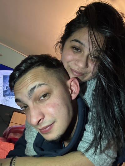 The image displays two adult individuals, one male and one female, positioned in close proximity within an indoor environment. The male is in the foreground, facing the camera with a direct gaze. He possesses dark hair styled with a fade, distinct eyebrows with a visible cut or shave mark on the left, light brown eyes, and slight stubble on his chin. Redness is observed on his left cheek. He is wearing a dark blue hooded sweatshirt with a grey internal hood lining. A multi-strand beaded bracelet is present on his left wrist. The female is positioned behind the male, leaning against him with her head resting near his left shoulder. She has long dark hair, dark eyes, and a faint smile directed towards the camera. She appears to be wearing a grey garment. The background is out of focus but reveals a television screen or monitor on the left, displaying text and graphical content, and a reddish-orange textile object in the lower left quadrant. The ceiling is a light, warm hue, suggesting artificial interior illumination. The specific location within the city of Kastel, Bulgaria, cannot be determined from the visual information provided.