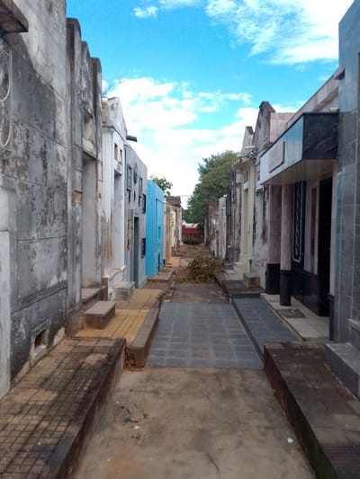 This image depicts a narrow, weathered alleyway within the Recoleta Cemetery in Asuncion, Paraguay. Tall, stone mausoleums line both sides of the alley, showing signs of age and disrepair with discolored walls and visible wear. The pathway is uneven, composed of a mix of gray and yellow tiled sections interspersed with bare earth. Debris, including fallen branches, litters the path. In the background, a red structure is partially visible. The sky above is bright blue with scattered white clouds. There are no people visible.