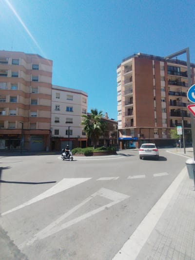 The image depicts a street scene in Rubí, Spain, featuring a roundabout with a circular green area in the center containing a palm tree. On the left side of the street, there is a beige and pink building with multiple floors, with commercial establishments on the ground floor, including a "Tabacs" store. A person on a scooter is seen driving around the roundabout. A white car is also present, driving on the roundabout's opposite side. On the right side, a tall brick building with balconies is visible. Road markings, including white directional arrows, are painted on the street. A trash receptacle and various road signs are visible on the right side of the scene. The sky is clear and blue.