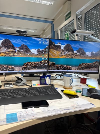 The image shows a desk in an office environment. There are two computer monitors displaying a mountain and lake landscape. A webcam is mounted on top of the left monitor. A keyboard, a mouse, a calculator, a hole punch, and a mug are on the desk. There are also some papers, sticky notes, and pens. In the background, a window with blinds and an emergency exit sign are visible. A sign that reads "Notausstieg" is also present. The office appears to be a standard workspace. There is no visible indication that this scene is located in Mannheim, Germany.