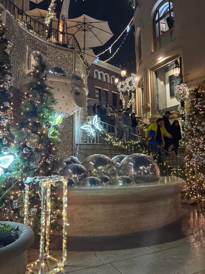 A nighttime scene features a fountain adorned with shiny silver spheres, and Christmas decorations including lit trees and oversized ornaments. People are seated on a bench under festive lighting. The backdrop includes buildings with illuminated windows and architectural details. A white awning is visible on the left side of the frame. The overall impression is of a lively, decorated urban setting.