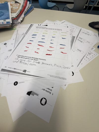 The image depicts a close-up, top-down view of a desk littered with papers and other stationery. The primary focus is on a worksheet with a grid of words, some of which are highlighted in red and yellow. Below this worksheet, a handwritten message in German reads: "Ich esse gerne in der Stadt Pommes, Pizza, Döner und Brezeln." This translates to "I like to eat fries, pizza, doner, and pretzels in the city." Another worksheet visible in the lower right corner is from "Verlag an der Ruhr | Autorin: Bogumila Baumgartner ISBN 978-3-8346-3551-8 | www.verlagsruhr.de." This information indicates the materials are likely from a German educational publisher. The presence of "Dorsten" is not explicitly mentioned in the image itself, but "Verlag an der Ruhr" implies a connection to the Ruhr region of Germany, where Dorsten is located. The overall scene suggests a classroom or study environment, with papers, pens, and other learning materials spread out.
