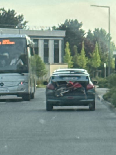 The image depicts a street scene in Villebon-sur-Yvette, France. A bus is driving on the right side of the road, followed by a dark-colored car. A bicycle appears to be partially inside the trunk of the car. Trees and buildings line the street. A tall pole is visible on the left side of the image.